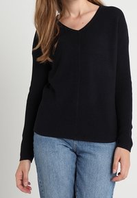 Schwarzer, gerippter Pullover mit V-Ausschnitt und langen Ärmeln, mit strukturiertem Design und lockerer Passform. Kombiniert mit hellblauen Jeans.
