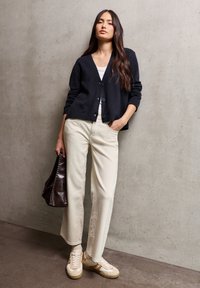 Granatowy sweter z dzianiny z guzikami, dopasowany do białej bluzki, w zestawie z beżowymi spodniami o szerokich nogawkach i beżowymi trampkami, trzymając ciemnobrązową torbę.