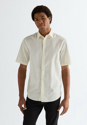 Koszula na krótki rękaw na guziki w jasnym kremowym kolorze, z fakturowanej tkaniny, z klasycznym kołnierzem i jasnymi guzikami, zestawiona z ciemnymi spodniami dżinsowymi.