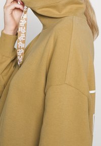 Senapsgul oversized hoodie med hög krage, kontrasterande vit detalj och mönstrad dragsko. Mjuk bomullstyg med ribbade ärmslut.