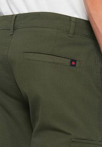 Grüne Baumwollhose mit einer flachen Gesäßtasche und einem Logo-Detail in Marineblau und Rot. Der Stoff hat eine subtile texturierte Oberfläche.