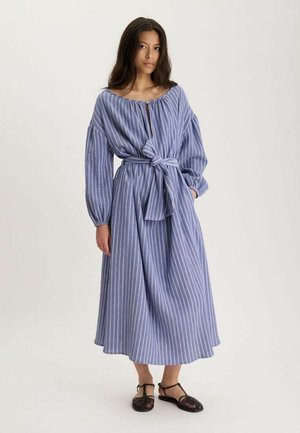 Femme portant une robe midi à manches longues rayée bleu et blanc avec une taille nouée, associée à des sandales plates tressées noires.