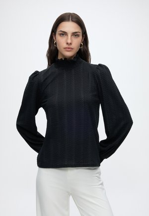 Junge Frau mit dunklen, glatten Haaren, die goldene Creolen, eine strukturierte schwarze Langarmbluse und weiße Hosen vor einem schlichten Hintergrund trägt.
