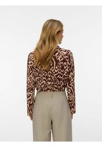 Femme aux longs cheveux blonds portant un haut à manches longues à motifs marron et beige, rentré dans un pantalon beige taille haute, vue de dos.