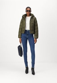 Giacca piumino verde oliva, taglio strutturato, con cappuccio. Camicia bianca, jeans skinny blu e stivaletti neri con punta. Porta una borsa blu navy.