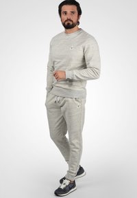 Helles graues Sweatshirt und passende Jogginghose aus weichem Material; mit kleinem Logo auf der Brust und elastischen Bündchen. Trägt dazu dunkelgraue Sneaker.