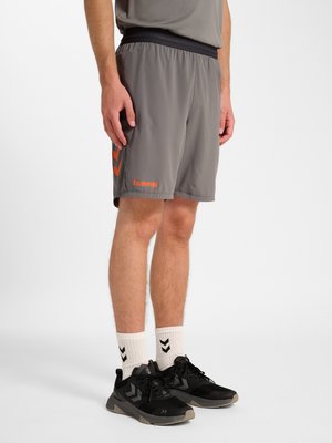 Mann trägt graue Hummel-Shorts, weiße Socken mit schwarzem Chevron und schwarze Sportschuhe, steht vor einem einfarbigen Hintergrund.