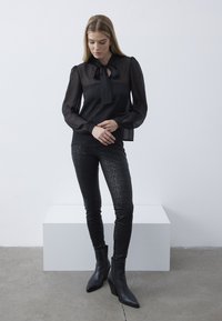 Zwarte doorschijnende blouse met een strikdetail bij de hals, gecombineerd met aansluitende zwarte broek met een gestructureerd bloemenpatroon en puntige enkellaarsjes.