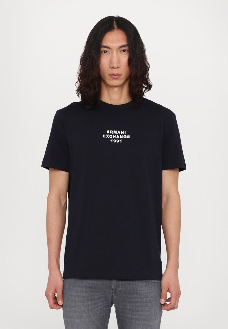 T-shirt en coton bleu marine avec un logo blanc "Armani Exchange 1991" sur la poitrine, col rond et manches courtes, porté avec un jean gris.