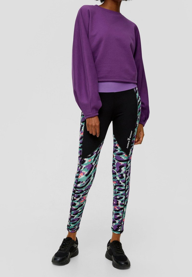 Sudadera morada con mangas anchas, combinada con leggings negros que presentan un patrón abstracto colorido y un logo. Zapatillas negras completan el look.