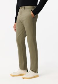 Khaki slim-fit broek met een gladde textuur, riemlussen en zijzakken, gecombineerd met witte sneakers met een groene accent.