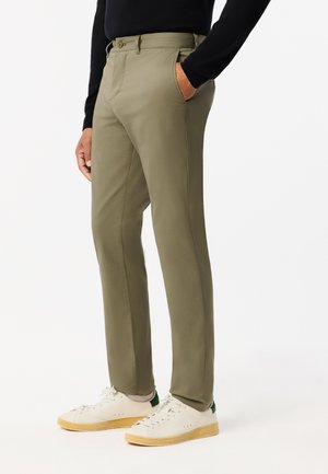 Khaki slim-fit broek met een gladde textuur, riemlussen en zijzakken, gecombineerd met witte sneakers met een groene accent.