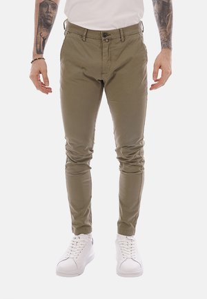 Uomo con pantaloni verde oliva slim fit, scarpe da ginnastica bianche, camicia bianca e braccialetti, con avambracci tatuati visibili, in piedi davanti a uno sfondo semplice.