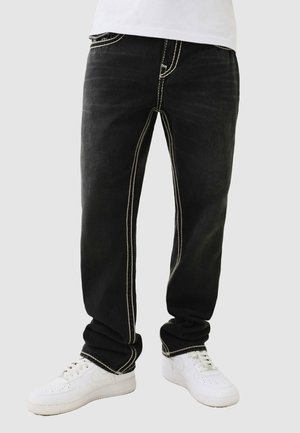 Parte inferiore del corpo con jeans neri con cuciture bianche e sneaker bianche su uno sfondo neutro.