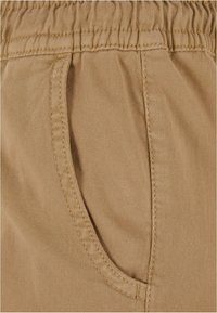 Gros plan sur la poche d'un pantalon en tissu beige avec des coutures visibles et un détail de ceinture élastique.