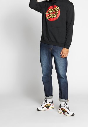 Haut à capuche noir avec le logo "Santa Cruz" rouge et jaune, associé à un jean en denim bleu et des baskets blanches avec des accents noirs et jaunes.