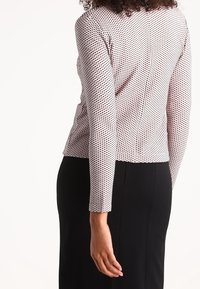 Haut à manches longues de couleur rose clair avec un motif à pois noirs, design ajusté et texture lisse, associé à une jupe sombre.