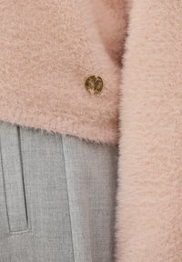 Cardigan rose pâle à texture douce avec un petit bouton rond doré portant un logo, sur un vêtement en tissu plissé gris.