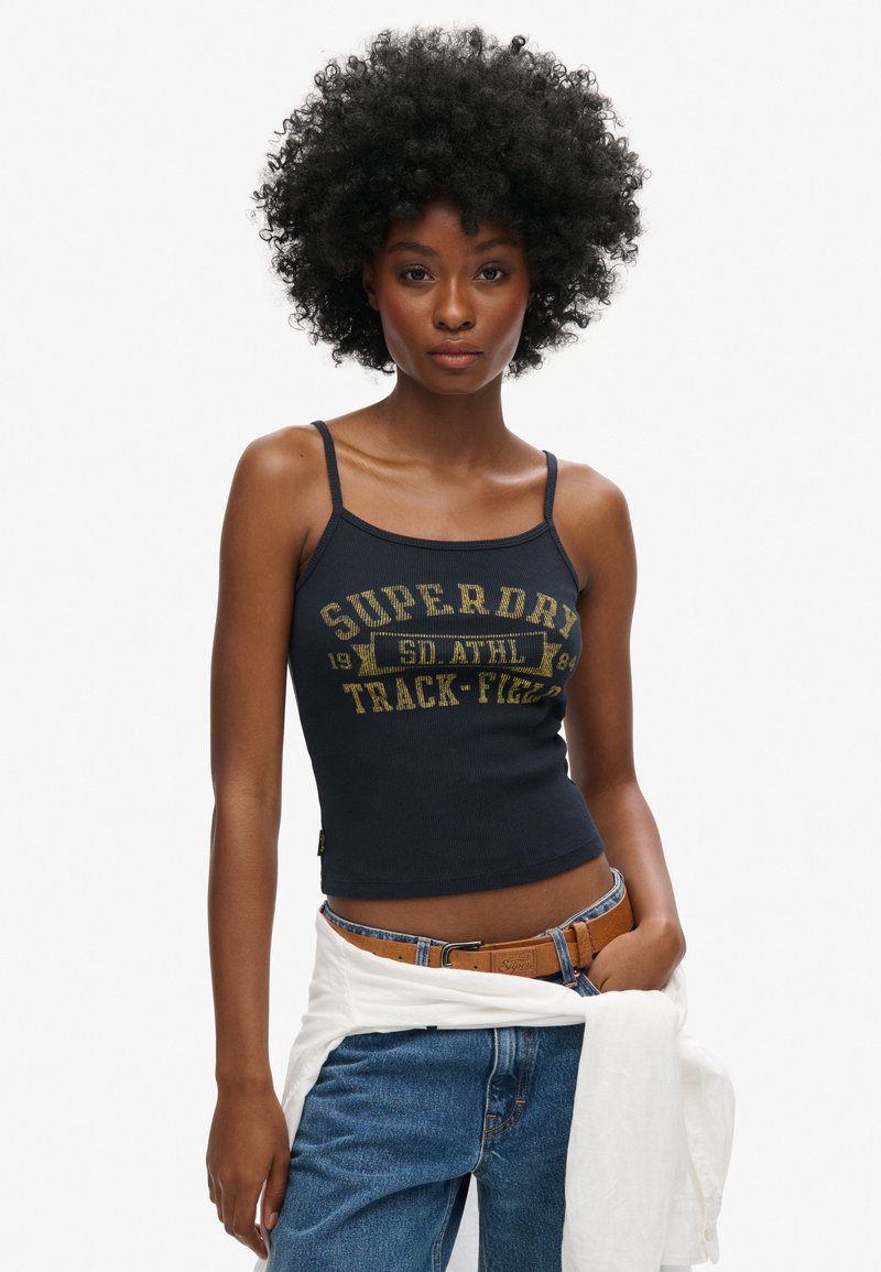 Débardeur côtelé bleu marine avec un graphique "Superdry" jaune ; design ajusté avec des bretelles fines, associé à un jean en denim bleu et une chemise blanche nouée à la taille.