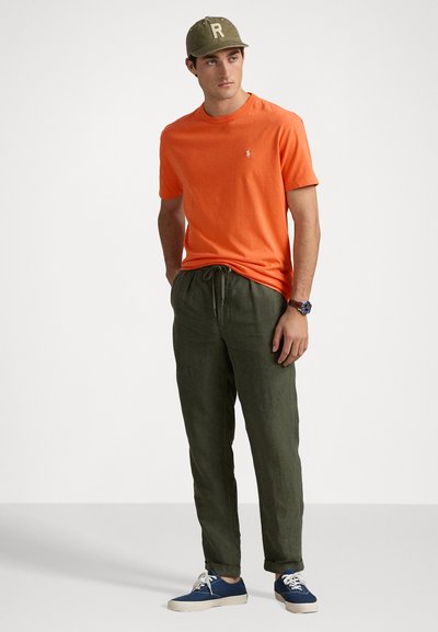 Orange kortärmad T-shirt med en liten logotyp, kombinerad med gröna linnetrousers med dragsko och blå canvas sneakers. En keps med bokstaven "R" ingår.