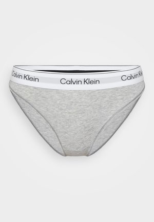 Szare slipy Calvin Klein z białym elastycznym paskiem w talii, na którym znajduje się czarny logo Calvin Klein, na jednolitym jasnym tle.