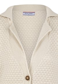 Beige kardigan s texturovaným pleteným designem, s klopovým límcem a jedním hnědým knoflíkem vpředu. Na vnitřní straně je viditelný štítek značky.