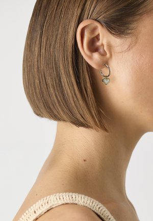 Orecchini a cerchio dorati con un piccolo charm a forma di cuore, caratterizzati da una finitura liscia e un design delicato, indossati su un orecchio da una persona con capelli corti.