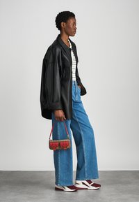 Μαύρο δερμάτινο υπερμεγέθες σακάκι, λευκή μπλούζα με ρίγες, φαρδιά μπλε τζιν, κόκκινη τσάντα χειρός με σχέδια και μπορντώ αθλητικά παπούτσια.