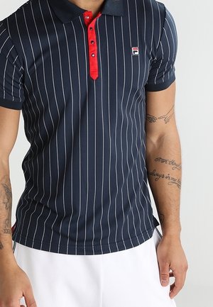 Polo bleu marine à fines rayures blanches verticales, patte de boutonnage rouge et petit logo sur la poitrine, porté par un homme tatoué avec un short blanc.