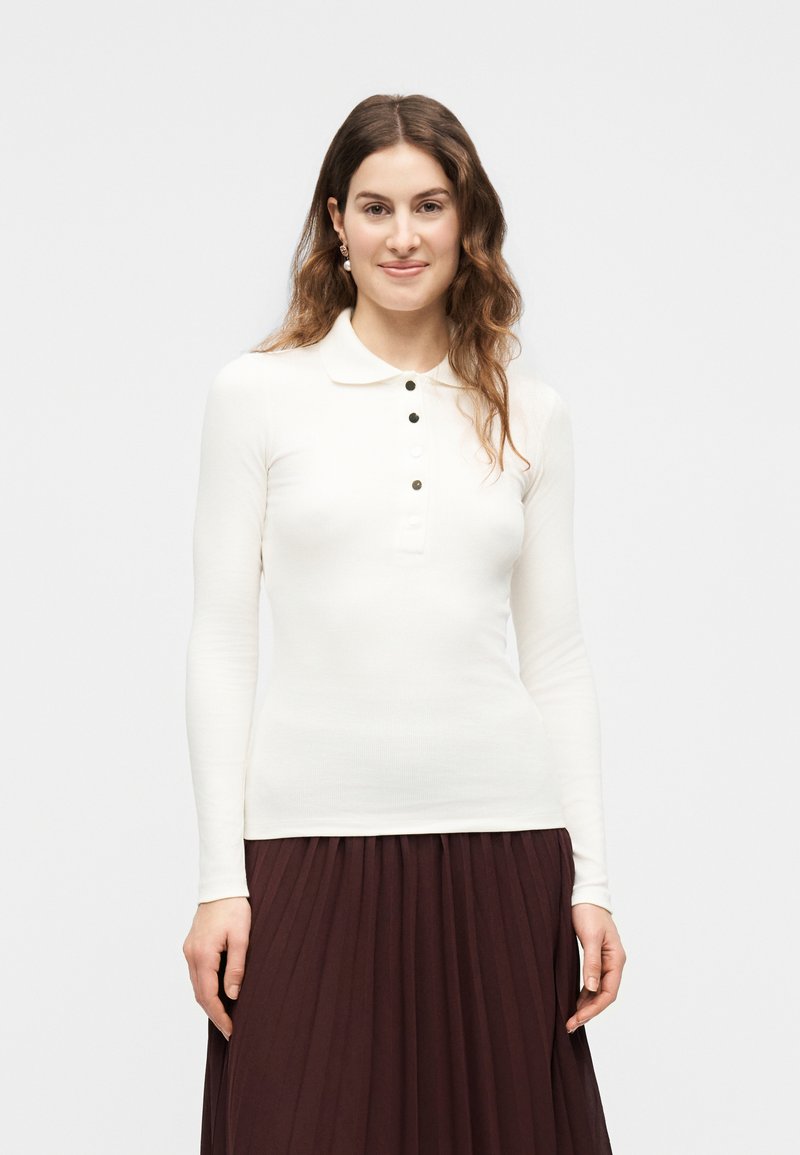 Γυναίκα με κυματιστά καστανά μαλλιά φοράει λευκό πόλο με μακριά μανίκια και σκούρα πλισέ φούστα, χαμογελώντας μπροστά σε απλό φόντο.