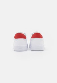 Sneakers bianche con accenti rossi sul tallone, texture in pelle liscia, design a basse e dettagli del logo sul tallone. Aspetto semplice e moderno.