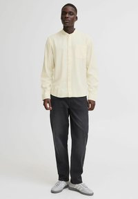 Camisa de manga larga, amarillo pálido, con botones en la parte frontal y un bolsillo en el pecho, combinada con pantalones gris oscuro y zapatillas grises. Textura suave.