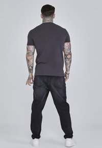 Donkergrijze t-shirt met korte mouwen, gecombineerd met zwarte jeans, van achteraf gezien. Bevat tattoos op de armen en witte sneakers.