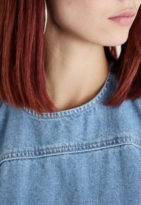 Denim krekls ar apaļu kakla izgriezumu, ar redzamām šuvēm un gaiši zilu krāsu. Modelim ir sarkani mati un lieli sudraba auskari cilpās.