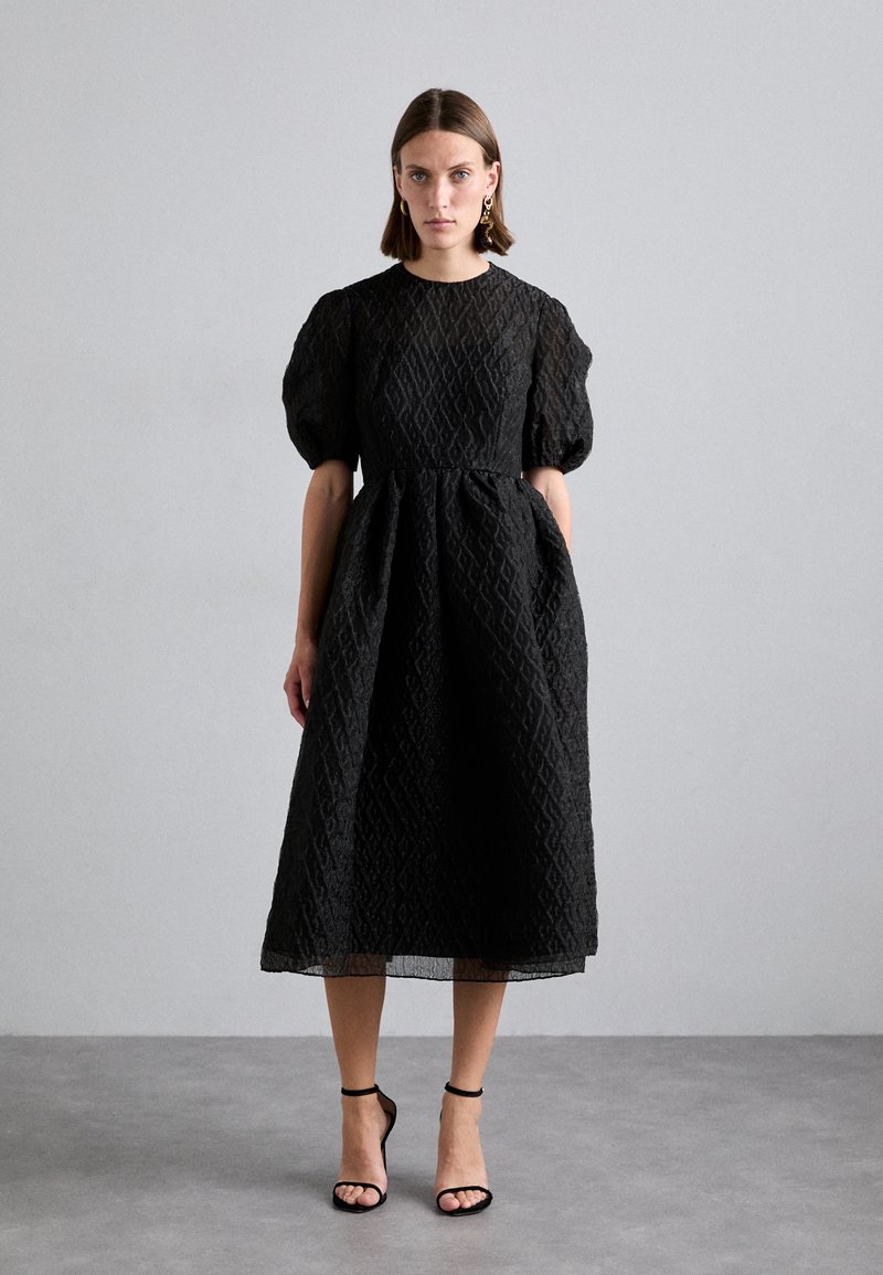 Black puff-sleeve dress in textured fabric with a diamond pattern, cinched waist, and knee-length skirt. Paired with black strappy heels.