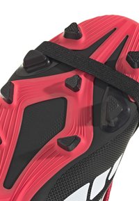 Κοντινή όψη της σόλας ποδοσφαιρικού παπουτσιού κόκκινου και μαύρου χρώματος με τριγωνικούς καρφίτσες και μαύρο λουράκι στο μέσο.