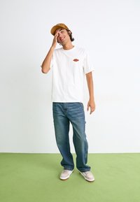 Hvid bomulds t-shirt med et lille orange logo, parret med blå denim jeans og beige sneakers. Baggrunden har et grønt gulv.