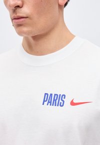 Κοντινό πλάνο στον λαιμό και το στήθος ενός ατόμου που φορά άσπρο μπλουζάκι με τη λέξη «PARIS» με μπλε γράμματα και το κόκκινο λογότυπο Nike Swoosh.