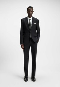 Completo nero con un design aderente, camicia bianca, cravatta a fantasia in bianco e nero, fazzoletto bianco e scarpe nere.
