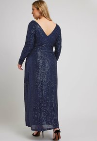 Abito lungo blu navy con dettagli in paillettes, scollatura a V profonda sul retro e maniche lunghe. Presenta una gonna lunga fino al pavimento e uno spacco laterale per il movimento.