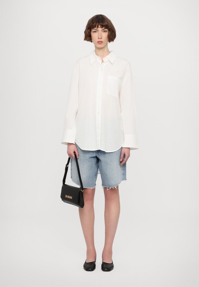 Camicia bianca con bottone e tasca, abbinata a pantaloni shorts in denim chiaro con bordi sfilacciati. Borsa nera con lettering dorato, scarpe nere piatte.