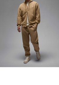 Sudadera con cremallera color beige con bolsillos tipo canguro, pantalones de chándal beige a juego y zapatillas de deporte de color claro, con un logo en el pecho y en los pantalones.