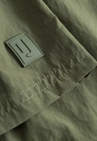 Olajzöld szövet sima tapintással, egy téglalap alakú, "J" betűvel emboszolt rugalmas címkével. Az illesztéseknél láthatóak a varrások.