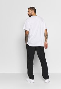 Homem com tatuagens a usar uma t-shirt branca, calças pretas e ténis brancos está de costas contra um fundo claro e liso.