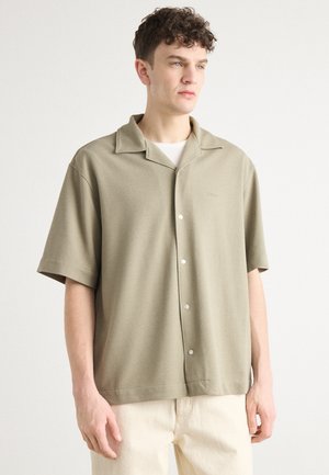 Camicia a maniche corte con colletto di colore verde oliva, realizzata in tessuto testurizzato, con chiusura frontale a bottoni e dettagli con logo in rilievo.
