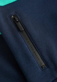Marine tekstil med en glat tekstur har en lynlås-lomme og en mærket læderlap med "BAKER BY TED BAKER" prægede.