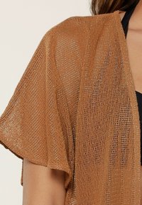 Kimono aperto di colore marrone, realizzato con un tessuto a maglia aperta, caratterizzato da un motivo testurizzato e fili metallici. Maniche corte e vestibilità rilassata.