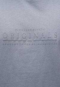 Γκρι ύφασμα με ανάγλυφο κείμενο που αναγράφει "Nineteen Ninety Originals 1990 Brought to you by Jack & Jones" στο κέντρο.