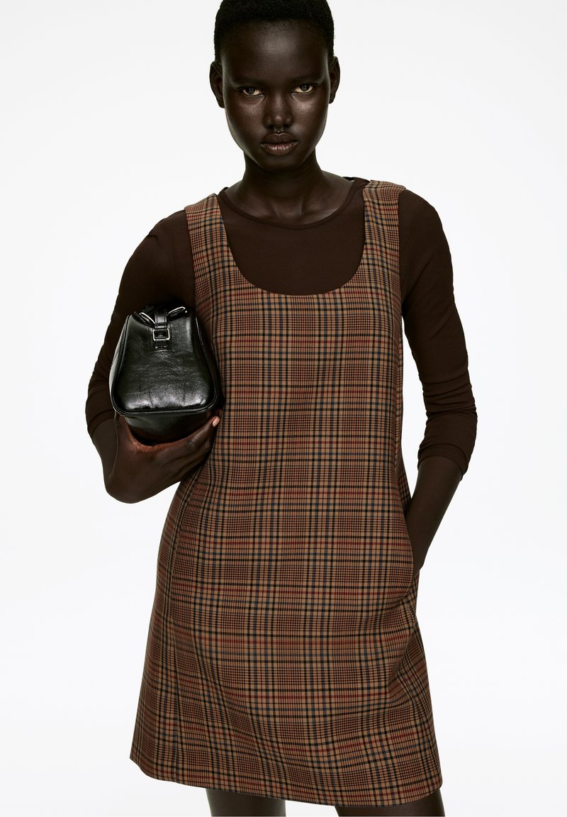 Braun karierter Kleid mit Rundhalsausschnitt, aus einem Stoff mit glatter Textur. Model hält eine kleine schwarze Lederhandtasche.
