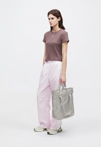 Γυναίκα ντυμένη με μωβ τεχνικό μπλουζάκι Nike, ανοιχτό ροζ φόρμες, μπεζ αθλητικά παπούτσια, κρατώντας μια γκρι τσάντα tote Nike, στέκεται μπροστά από ένα λευκό φόντο.